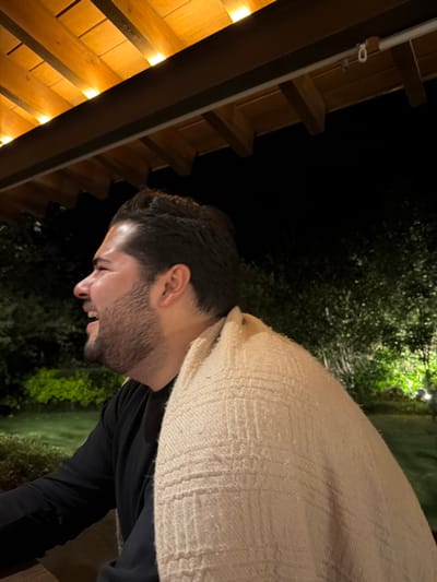 The image displays an adult male, captured from the chest up in profile, facing left, in an outdoor or semi-outdoor setting at night. The male has dark hair and a beard, and is wearing a dark, long-sleeved shirt. A light-beige, woven blanket with a textured grid pattern is draped over his right shoulder and back. His mouth is open in an expression indicating laughter or a broad smile, with his head tilted slightly backward. Above the subject, a wooden structure, likely a pergola or roof section, is visible, featuring integrated warm-toned artificial lights. The background consists of dark foliage, which is intermittently illuminated by green-toned ground lights, suggesting a landscaped area or garden. The specific location within Valle de Bravo, Mexico, is not identifiable from the visual information presented. The scene is illuminated by artificial light sources.