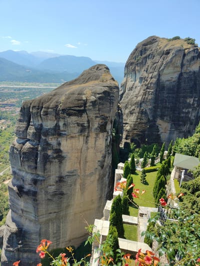 The image captures a scenic view of the Meteora rock formations in Kalambaka, Greece, under a clear blue sky. Towering, sheer rock cliffs dominate the foreground and midground, displaying textured surfaces with variations in light and shadow. In the midground and right side of the image, a well-maintained garden area is visible. This garden features neatly trimmed cypress trees, a manicured lawn, and a small gazebo with a green roof. A stone pathway and low stone walls delineate the garden spaces. Orange trumpet-shaped flowers bloom in the foreground and along the edge of the garden, adding a splash of color to the scene. The background reveals a hazy vista of rolling hills and distant mountains, with a glimpse of the town of Kalambaka nestled in the valley below. The scene is devoid of any people.