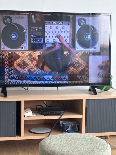 A flat screen television is mounted to the wall in a living room in Schaerbeek, Belgium. The TV is displaying a video of a woman with long, dark hair, wearing a black tank top, adjusting a sound mixer that is connected to two turntables. The turntables are on a wooden table covered in a patterned rug with geometric shapes in red, black, and tan hues. The scene in the TV is illuminated by overhead lighting. The TV is resting on a wooden TV stand, which has two shelves, a black cabinet door, and a light brown top. The top shelf of the TV stand is holding a black box, a white rectangular object, and white wires. The bottom shelf holds a round object with a white label, a blue and white box, and black wires. An out-of-focus, light green ottoman sits in front of the TV stand.