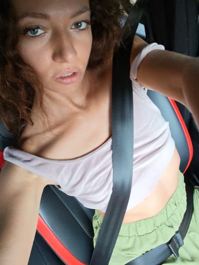 A woman with curly brown hair and green eyes is in a car, wearing a pale pink tank top and green shorts, with a seatbelt across her chest. The car has black seats with red accents. The location is implied to be Minh Tân Commune, Vietnam.