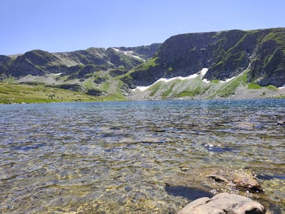 This image depicts a serene natural landscape at the Seven Rila Lakes in Panichishte, Bulgaria. The foreground features the clear, shallow water of a lake, revealing the rocky bottom beneath the surface. Sunlight glints off the ripples, creating a shimmering effect. In the mid-ground, the lake extends towards a grassy shore where several people are visible, appearing to be walking or resting. Beyond the shore, the terrain rises sharply into imposing, green-covered mountains. Patches of snow are visible in the crevices and higher elevations of the mountains, indicating a cool climate even in summer. The sky is a clear, bright blue, with no clouds visible. The overall impression is one of a tranquil, mountainous environment, likely a popular destination for hikers and nature enthusiasts.
