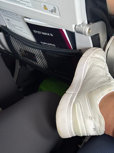 A white sneaker rests on the edge of an airplane seat pocket containing a safety manual labeled "B737 MAX 8 Instructivo." The seat's metallic armrest is visible to the left. A leg covered in black leggings is to the right of the seat pocket and under the sneaker.  The sneaker appears dirty, and the seat pocket is made of black mesh.