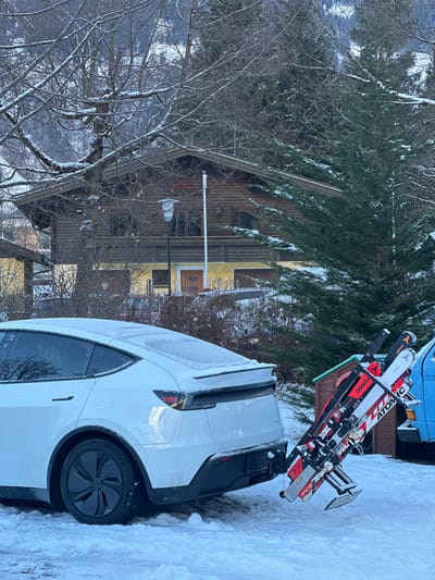 A snow-dusted white car parks in a snowy area next to a house. Skis are mounted on a carrier attached to the car's rear. A mix of bare trees and snow-covered evergreens surround the scene. The house has a traditional design, partially obscured by trees. A light pole stands in front of the house. The weather appears cold and overcast.