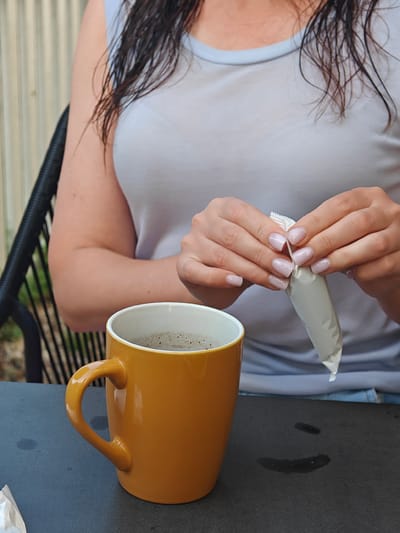 The image captures a close-up view of a person's hands opening a small, white packet over a dark surface. The person is wearing a light blue tank top and has dark, wet hair that falls around their shoulders. Their fingernails are neatly manicured with a pale, shimmery pink polish.
In the foreground, to the left, sits a vibrant orange mug filled with a dark liquid, likely coffee. The mug has a prominent, curved handle and is positioned on the dark surface, which appears to be a table. A few dark spots are visible on the table, suggesting it might be slightly damp or stained.
The background shows a portion of a chair with a woven texture and a hint of greenery, indicating an outdoor setting. The lighting suggests it could be daytime, possibly a bit overcast. The overall mood is relaxed and casual, as the person prepares to add something from the packet to their drink. The scene is set in Étretat, France. There is no visible text in the image.