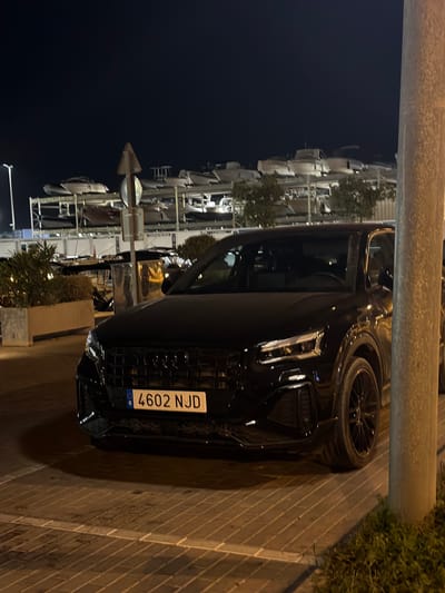 The image shows a black Audi Q2 parked on a paved surface at night in Ibiza, Spain. The car is positioned in the foreground, with its front end facing the viewer. The license plate, "4602 NJD", is clearly visible. Behind the car, a multi-level structure holds several boats on racks, suggesting a marina or boat storage area. The background is dark, indicating it is nighttime, with some distant lights visible. The scene is illuminated by artificial light sources, casting shadows on the ground. A lamppost is visible on the right side of the image. There are no people or any observable action in the scene.