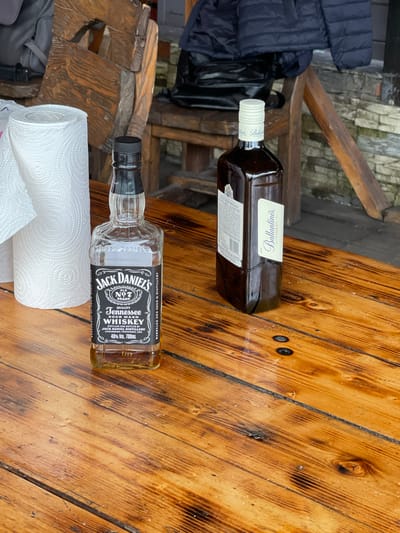 The image shows two bottles of alcohol lying on a wooden table, alongside a roll of paper towels. One bottle is Jack Daniel's Tennessee Whiskey, and the other is Ballantine's Finest Scotch Whisky. The setting appears to be outdoors, with a glimpse of a wooden structure and paving stones in the background. Based on context the location is possibly Картмазово, Russia.