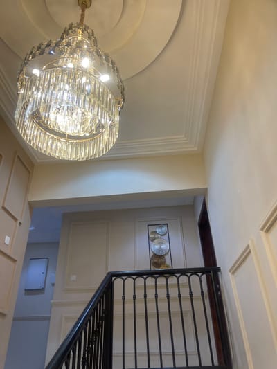 This interior photograph captures a luxurious and well-appointed space, likely within a home or commercial building in Adenta Municipality, Ghana. The scene is dominated by a large, ornate crystal chandelier suspended from a light-colored ceiling. The chandelier features multiple tiers of shimmering glass or crystal elements, brightly illuminated and serving as a central focal point.
To the right of the frame, a grand staircase with a black wrought-iron railing, showcasing decorative scrollwork, ascends. The railing is topped with a dark wooden handrail. The walls are painted in a light, neutral tone and feature elegant architectural details, including decorative paneling and intricate crown molding that extends across the ceiling. Around the base of the chandelier, the ceiling also exhibits a large circular molded design.
On a wall further back, beyond the staircase, a piece of modern wall art is displayed. It consists of multiple circular metallic discs – some silver, some gold – arranged within a dark rectangular frame. Above this artwork, there is a recessed opening or shelf with dark wooden trim. Below the visible staircase, a glimpse of a lower floor reveals a light-colored tiled or paneled surface and a white rectangular access panel or utility box mounted on a wall.
The photograph is taken in a well-lit interior, with the chandelier actively providing illumination. No people are visible, and there is no discernible activity taking place. The time of day and weather conditions cannot be determined from this indoor setting. No text is visible in the image.