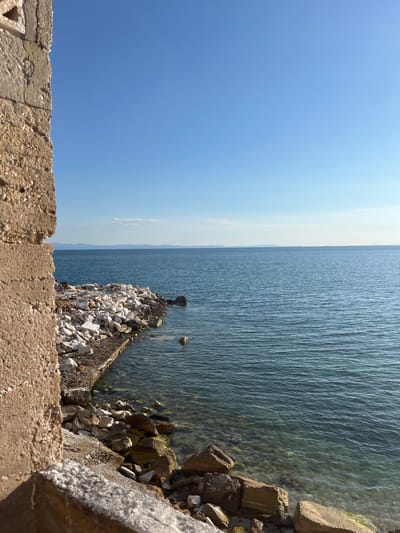 The image depicts a sunlit scene of the sea and coastline in Potos, Greece. In the foreground, a rough, textured concrete wall occupies the left portion of the frame, its weathered surface showing signs of age. Beyond the wall, a breakwater made of white and grey stones juts into the clear, blue-green water. The water is calm with gentle ripples, and the seabed is visible in shallower areas, littered with rocks. In the middle ground, a few people are visible near the stone breakwater, some sitting and others possibly standing, with what appears to be picnic items. The horizon is defined by a distant, faint mountain range under a clear, bright blue sky. The overall impression is one of a peaceful coastal landscape bathed in natural light.