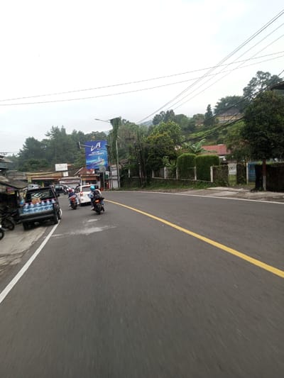 The image shows a street in a mountainous area, likely in Asia. The road is paved with a yellow line dividing the lanes. Several vehicles, including a small truck filled with water bottles and motorcycles, are present. A billboard advertising "LA Taste the Coolest Flavors" is visible. The surrounding environment includes trees, foliage, and houses on a hillside. The sky is overcast.