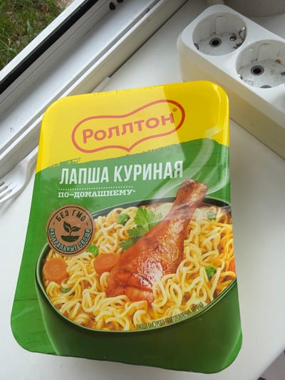 A package of Rollton chicken-flavored instant noodles is shown, with a close-up of the product and its packaging. The packaging is primarily yellow and green, with text in Russian. The image features a graphic of a chicken leg and noodles, with a circular "Bez GMO" (GMO-free) and "Naturalnye ovoshchi" (Natural vegetables) seal. The package is placed on a white surface, possibly a windowsill, with a white electrical outlet visible in the background. The scene is indoors, with some greenery visible through a window on the left.
