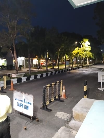 A nighttime exterior shot shows a road in Tambontunong, Indonesia, with streetlights illuminating a row of trees and buildings. A sign in the foreground reads "DILARANG KELUAR TANPA IZIN" (Forbidden to exit without permission). A person in a yellow shirt is partially visible on the left. Orange traffic cones and a portable barrier are present on the road.