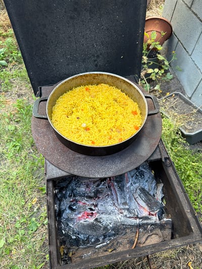 The image captures a close-up, overhead view of a meal being prepared outdoors. The main subjects are a large pot of yellow rice with visible pieces of carrot and possibly other ingredients, placed on a rustic metal outdoor cooking apparatus. Next to the pot, the apparatus contains glowing embers and charred wood, indicating a fire is burning.
The setting appears to be a backyard or garden area, with green grass and dry vegetation visible in the background. There are hints of a gray, textured surface, possibly concrete pavers, and a brown terracotta pot with a small, leafy green plant in the upper left corner. The overall scene suggests an outdoor cooking activity, possibly a barbecue or camp-style meal.
The time of day seems to be daytime, with sunlight casting shadows and highlighting the colors of the food and fire. The weather appears to be pleasant, suitable for outdoor activities. There are no people visible in the image. The focus is solely on the food preparation and the cooking environment. The image has a candid, unposed feel. No visible text is present in the image. The location context provided is Balivka, Ukraine.