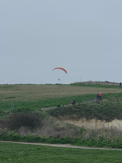 The image depicts an outdoor scene in the city of Thanet, United Kingdom, under a uniformly overcast, light grey sky. A single red paraglider, with a dark human figure suspended beneath its canopy, is airborne in the upper center of the frame. The terrain consists of multiple undulating green areas. In the immediate foreground, a paved pathway runs horizontally, bordered by short green grass interspersed with small white flowers. Above this path, a steep bank covered in dense green vegetation descends to an exposed, lighter-colored earth cliff face, possibly chalk, which then transitions into further green slopes. Three individuals are present on these slopes: one person, wearing a prominent red jacket, is positioned on a higher verdant slope towards the right, appearing to walk or stand. To their left and slightly lower, another individual in darker clothing is seated or crouching near a distinct red object on a green embankment. A third, distant figure is faintly visible near the horizon on the far right. A single dark, slender post protrudes vertically from the dense green vegetation on the mid-left side of the scene.