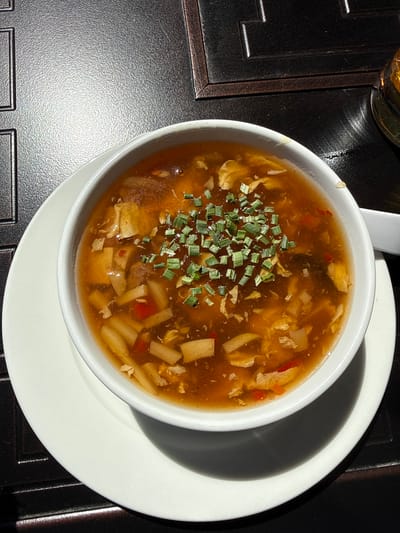 A close-up, high-angle shot captures a steaming bowl of soup, likely hot and sour soup, served on a dark, textured table surface. The soup, a rich amber-brown, fills a white ceramic bowl and is generously garnished with finely chopped green chives or scallions scattered across its surface.
Within the hearty broth, numerous ingredients are visible, including irregular pieces of shredded yellow egg, rectangular slices of light-colored bamboo shoots, and small cubes that appear to be tofu. Slender, bright red strips, possibly bell pepper or chili, add a touch of color to the mixture.
The white bowl rests on a slightly larger white plate or saucer, which casts a prominent, dark shadow across the table surface due to strong, direct lighting. A white ceramic spoon handle is partially visible at the top left, resting in the bowl. The table or placemat beneath is dark grey or black with a distinctive, slightly pebbled texture, and features a subtle raised pattern along its visible edges. In the upper left corner, a portion of a glass containing a golden-brown liquid, possibly beer or tea, is partially visible.
The scene is brightly lit by what appears to be direct sunlight, casting sharp, defined shadows and highlighting the texture of the table surface, suggesting a daytime setting, possibly outdoors or near a window in Hainburg an der Donau, Austria. No people or visible text are present in the image.