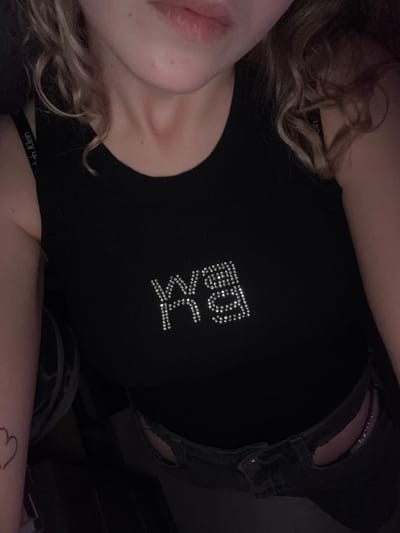 A selfie-style, upward-angled shot captures a person wearing a black top with "BK" embellished in rhinestones on the front. The person's face is partially visible, showing lips and a hint of a cheek. Blonde, curly hair frames their face. The black top appears to be layered over a Calvin Klein-branded undergarment, as the straps are visible. The person is also wearing dark jeans with a rip near the waistband. A small tattoo of a heart is visible on the arm. The overall scene is darkly lit. Turin, Italy, cannot be inferred from the image.