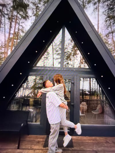 A young couple is embracing on the wooden deck of an A-frame cabin in the woods. The man, wearing a light-colored tracksuit and white sneakers, is holding the woman, who is also dressed in a matching tracksuit and white sneakers. They are looking at each other, and the woman's legs are wrapped around the man's waist. The cabin has large glass windows that offer a view of the surrounding forest with tall trees. The setting is rustic and outdoorsy. The location is Beșalma, Moldova.