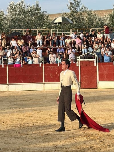 In an outdoor arena in San José del Valle, Spain, a matador is walking, facing right, holding a red cape. He is dressed in a light-colored cropped jacket and dark pants tucked into black boots. Behind him, a red wall partially obscures a crowd of spectators seated in stands. In the background, trees and a light sky are visible.