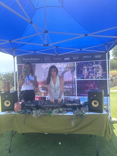 The scene depicts an outdoor event, possibly in Pueblo Nuevo de Jasso, Mexico, identifiable from the signage of "VidantaWorld Mexico Open." Under a blue canopy structure, a DJ setup is arranged on a table covered with a light green cloth. The setup includes two black KRK speakers, a Pioneer DJ mixer, and associated wires. The setup is adorned with floral decorations. Two women are visible in the scene. One is using a smartphone to take a picture, while the other is operating the DJ mixer. The background features promotional images with the VidantaWorld logo, advertising various attractions like "Cirque du Soleil Joya" and "JUNGALA AQUA EXPERIENCE." The event seems to be taking place on a grass surface.