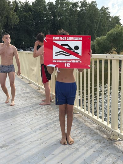 The image shows three young men on a concrete walkway next to a body of water, likely a river or lake, under a bright sky. The perspective is from above, looking down.
The two men in the foreground are shirtless and wearing swim trunks. The man at the bottom is facing away from the camera, showing his patterned swim trunks. The man above him is also facing away, wearing black and red swim trunks. He is looking back and appears to be gesturing or speaking.
A third young man, in the middle ground, is holding up a red sign with a white prohibition symbol on it. The symbol depicts a person swimming, crossed out. The sign has Russian text. The man holding the sign is shirtless and wearing blue swim trunks. He is looking down at the sign.
The walkway has a light-colored surface, possibly concrete or stone, with some debris scattered on it. A metal railing runs along the edge of the walkway, separating it from the water. The water is rippling and reflects the sunlight. Trees with green foliage are visible in the background, suggesting a natural or park-like setting. The sun appears to be high in the sky, indicating it's daytime.
The visible text on the sign reads "КУПАТЬСЯ ЗАПРЕЩЕНО" which translates to "SWIMMING IS PROHIBITED". Below that, it says "ПРИ ПРОИСШЕСТВИИ ЗВОНИТЕ: 112", meaning "IN CASE OF EMERGENCY CALL: 112". The presence of this sign and the men in swim trunks suggest that swimming is not permitted in this area, despite the proximity to the water. There is no clear indication of distress or urgency among the men.