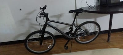 A gray Huffy mountain bicycle is indoors, leaning against a white wall next to a small round black table. The bicycle has black tires, a black seat, and a kickstand. The pedals are black, and the frame has the word "HUFFY" printed on it. The table has a dark surface and a single leg. The floor is wooden, and a light-colored rug is partially visible in the foreground. There are electrical cords and a black object (likely an electronic device) on the table. The scene is likely in Fort Smith, United States of America, but there are no specific landmarks or identifiers to confirm.
