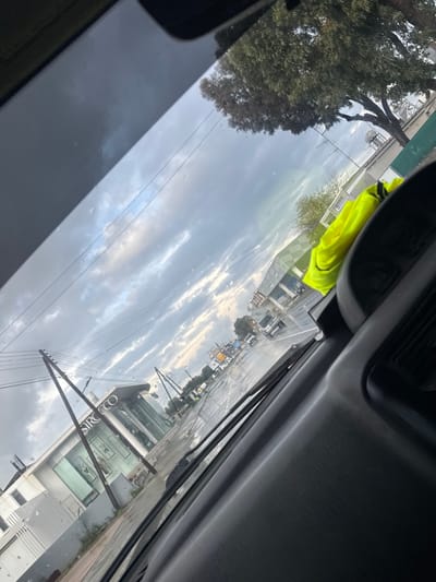The image displays a street view from inside a vehicle, looking through a windshield with minor water streaks, in Kato Polemidia Municipality, Cyprus. The foreground includes a dark-colored vehicle dashboard, with a fluorescent yellow high-visibility vest resting on the dashboard to the right.

The scene features a wet asphalt road, indicating recent precipitation. On the left side of the road, a modern commercial building with white paneling and large glass windows is visible, featuring a prominent "SIROCCO" sign. Multiple utility poles with overhead wires line this side of the road. Further down the street, on the right, other commercial buildings are present, including one with a distinctive green and grey checkered pattern on its upper facade. Several vehicles, including a light-colored van, are observed in motion on the wet road in the mid-distance. The sky is overcast with varied shades of grey clouds, and a large tree with dense green foliage is visible on the upper right, behind the street infrastructure.