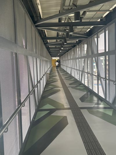A long, modern pedestrian walkway or enclosed bridge is depicted, extending into the distance with a strong sense of perspective. The structure is primarily composed of gray metal, featuring a perforated mesh floor and ceiling panels that allow light to filter through, creating distinct patterns of light and shadow.
On the left side, the walkway is supported by a robust metal framework of beams and diagonal struts. Several cylindrical spotlights are mounted along this wall, suggesting artificial illumination is also possible. Pipes are visible underneath the walkway structure on this side. The right wall is solid, adorned with a contemporary geometric mural in shades of green, gray, and white. A textured, rectangular strip, possibly a tactile warning or design element, runs horizontally across this patterned wall. A metal handrail runs along the right side of the walkway, attached by small hooks to the ceiling.
At the very far end of the walkway, a bright yellow structure or area is visible. A person, appearing small due to the distance, is walking away from the viewer in that direction. Near the end, yellow and black striped warning tape and a white box are placed on the floor. The overall setting suggests a public or commercial urban environment, such as the Subang Jaya City Council in Malaysia, designed for pedestrian traffic. The natural light filtering through indicates it is daytime. No distinct, readable text is prominently visible.
