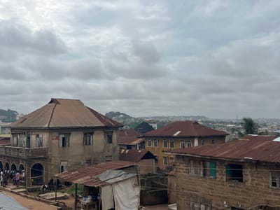 The image depicts a densely populated urban area in Abeokuta, Nigeria, characterized by traditional architecture and a cloudy sky. The foreground is dominated by a cluster of buildings with corrugated iron roofs, some showing signs of rust and age. One prominent building on the left features a multi-story facade with balconies and arched openings on the ground floor, where several people are gathered. These individuals appear to be engaged in casual conversation or resting.

Further into the image, more buildings with similar tin roofs are visible, interspersed with other structures. The roofs vary in condition, with some appearing newer and others quite weathered. The overall impression is one of a lived-in, developing urban landscape.

In the background, a more distant cityscape unfolds, with buildings of varying heights and styles. The terrain rises in the far distance, with hills covered in vegetation, suggesting a natural landscape surrounding the urban sprawl. The atmosphere appears overcast, with diffused light illuminating the scene. The environment seems to be a mix of residential and commercial spaces, with elements of everyday life playing out on the streets and around the buildings.