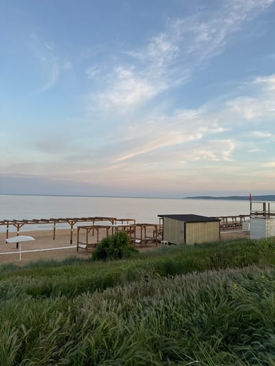 The image shows a beach in Береговой сельский совет, Ukraine. The view is from the shoreline, looking out over the water towards a distant landmass. The sky is light blue with scattered white clouds. On the right, there is a grassy area with tall, green vegetation. Wooden structures, possibly cabanas or shelters, line the beach, with some being covered and others open. The beach itself is sandy. The water is calm and reflects the sky's colors. A red flag is visible in the distance.