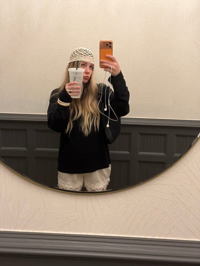 The media file shows a young woman taking a mirror selfie. She is wearing a black hoodie, a white crochet-style beanie, and white lace-trimmed shorts. She is holding an orange smartphone in her left hand and a clear plastic cup with a green logo in her right hand. White earbuds are plugged into her phone. Her blonde hair is long and wavy. The background appears to be a room with textured wallpaper and a large, dark-paneled door reflected in the mirror. The mirror is oval-shaped with a thin gold frame. The lighting suggests it is daytime indoors. The overall mood is casual and everyday. No visible text is present in the image, apart from the logo on the cup, which appears to be Starbucks.
