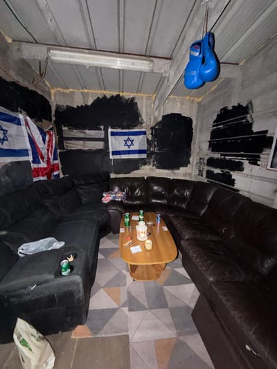 The image shows the interior of a room. The walls appear to be concrete or corrugated metal, with portions painted black. On the left wall, blue boxing gloves hang from a wire. Three flags are visible: one flag of Israel is mounted on the wall, and two flags (one being the UK flag and another being the flag of Israel) are on the floor. A dark leather couch is present in the room, along with a wooden table. Various items sit on the table, including what appears to be drinks and small objects. A rug with a geometric pattern is under the table. Debris and trash bags are near the couch, as well as a couple of beer cans inside the side of the couch. There is no indication in the image that this room is located in Yokneam Illit, Israel. Therefore, the location cannot be stated with confidence.