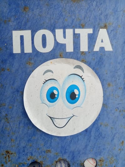 The image depicts a weathered blue surface with rust spots. On the left side, the white, vertically oriented word "ПОЧТА" (meaning "post" or "mail" in Russian) is visible. To the right of the word is a circular sticker featuring a cartoon face with large blue eyes, eyelashes, and a smile. The sticker has some minor damage. The location in the city of совхоза "Воронежский", Russia is not ascertainable from this image alone.