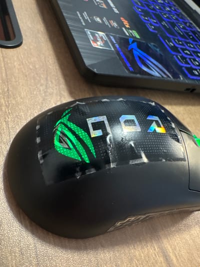This image shows a close-up of a black gaming mouse adorned with stickers. The stickers feature the "ROG" logo and the Republic of Gamers eye emblem, both with a holographic, iridescent sheen that shifts in color with the light. The "ROG" letters are placed vertically on the mouse, with the "R" at the top, followed by "O" and "G" below. The ROG eye emblem is prominently displayed below the letters. Some parts of the stickers appear to be peeling or lifting, suggesting they have been applied for some time. The mouse is positioned on a light brown, textured surface, possibly a desk or table. To the left of the mouse, a portion of a laptop is visible. The laptop has a black casing and a keyboard with blue, backlit keys. A prominent blue ROG logo is also visible on the laptop's palm rest. The overall scene suggests a gaming setup or a workspace related to gaming. There are no people visible in the image. The lighting appears to be from a natural source, possibly daylight, casting soft shadows. No specific time of day or weather conditions can be determined. The visible text includes "ROG" on the stickers and on the laptop, as well as what appears to be "JOIN THE REPUBLIC" below the ROG logo on the laptop. There are also other smaller text elements on the laptop, some of which are partially obscured. The context provided states "Małkowo, Poland," which is a geographical location, but there are no specific visual cues in the image that confirm this location.