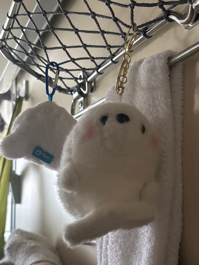 The image depicts an interior scene, characteristic of a train compartment, located in Kalachinsk, Russia. A white, round-bodied plush toy with black eyes and light pink cheek accents is centrally positioned, suspended by a gold-colored keyring from a polished metal rod. This rod forms part of an overhead storage system, which includes a tightly woven grey net directly above. To the left, a second, partially visible white plush toy hangs from a blue carabiner and displays a small light-blue tag with "PRIMUS" in white text. Below the main plush toy, a white terrycloth towel is folded and draped over a lower, parallel metal rail. The background consists of a plain, light-toned wall, with a vertical strip of green fabric or curtain visible on the far left side of the frame.