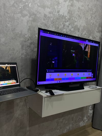 The image shows a room in Zubovo, Russia, with a gray wall and a white cabinet. A flat-screen LG TV is mounted on the wall. The TV is displaying a webpage with a movie and a chat interface with numbered emojis. A gray Acer laptop is connected to the TV, also displaying a webpage with emojis. A remote control is visible on top of the laptop. The scene suggests a person is watching a movie and interacting with others online.