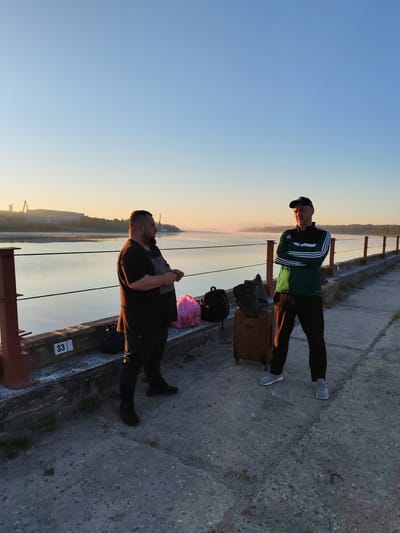 Two men stand on a concrete pier next to a body of water in Bol'shie Mkhi, Russia. The man on the left is heavier-set, wearing a dark t-shirt and dark pants. He is facing away from the camera, looking towards the water. Next to him are a pink bag and a black backpack. The man on the right is wearing a green and white tracksuit and a black baseball cap. He is facing the camera with his arms crossed. Beside him is a brown suitcase with wheels and a black backpack. In the background, there is a body of water with a thin layer of fog on its surface, and a distant industrial building. The sky is a clear blue with a hint of sunrise or sunset glow near the horizon. The pier has metal railings and concrete barriers. The pier has a number "33" marked on the barrier. The time of day appears to be either early morning or late evening, indicated by the soft lighting and the fog.
