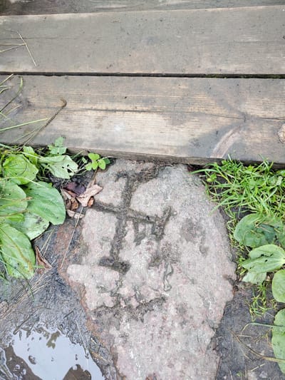 The image depicts a close-up view of a weathered stone with a symbol etched into its surface, located near a wooden walkway in Valaste, Estonia. The stone is partially covered by mud and surrounded by green vegetation and fallen leaves. Water is pooling on the ground next to the stone. The symbol appears to be a geometric shape, possibly a cross or a stylized letter, formed by dark lines on the lighter-colored stone. The wooden planks of the walkway are damp and appear to be part of an outdoor path, possibly in a park or natural area. The overall impression is one of age and natural erosion.