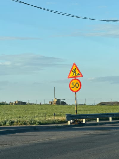 The image shows a road sign with a yellow triangle indicating road work, with a silhouette of a person shoveling. Below it, a circular sign indicates a speed limit of 50. The signs are mounted on a yellow and black striped pole, located next to a guardrail. In the background, there is a grassy field with some structures that appear to be old military vehicles or debris. The sky is blue with some clouds, and there are overhead wires visible at the top of the frame. The location appears to be a rural or semi-rural area, possibly on the outskirts of a city. There are no people visible in the image. The scene is lit by natural daylight, casting shadows on the road.