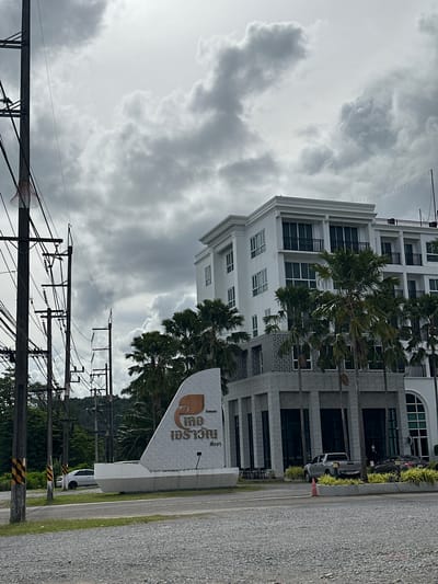The image captures a frontal view of a modern hotel, identified by a sign as "Chong Wang" in Thai, located in Phang-nga, Thailand. The hotel is a multi-story white building with dark-framed windows and balconies. In front of the hotel, palm trees are scattered. To the left of the hotel, a prominent white sign with Thai lettering and a logo is situated. The foreground is a gravel parking lot, and a paved road runs across the image. Utility poles with numerous wires are visible on the left side of the frame. The sky is heavily clouded with a dull, overcast appearance. Two vehicles, a silver pickup truck and a dark car, are parked in front of the hotel's entrance area. The environment appears to be tropical, given the palm trees and lush greenery in the background.