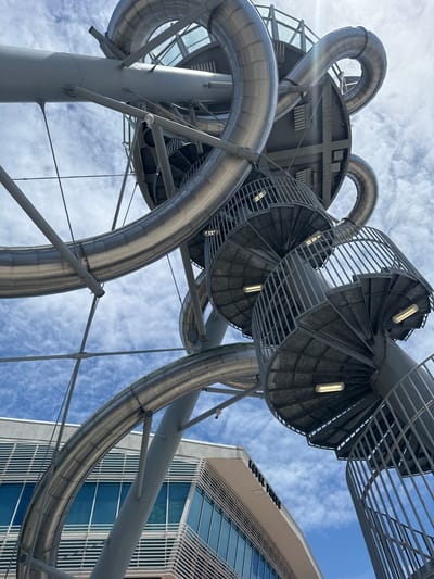 The image shows a low-angle view of the ArcelorMittal Orbit sculpture and slide in Queen Elizabeth Olympic Park, London, United Kingdom. The metallic structure features curved tubes and spiral staircases against a backdrop of a cloudy sky. A modern building with glass windows is visible in the lower left corner. There are no people or animals in the image. The scene is lit by daylight.