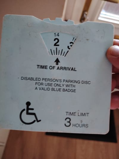 The image shows a close-up of a blue parking disc for disabled persons. The disc has markings for hours, with the number '2' and '3' clearly visible. An arrow points upwards to indicate the "TIME OF ARRIVAL". Below this, text reads "DISABLED PERSON'S PARKING DISC FOR USE ONLY WITH A VALID BLUE BADGE". A symbol of a person in a wheelchair is also present. At the bottom right, "TIME LIMIT" is written, followed by "3 HOURS". The disc appears to be held by a person's hand, with part of the person's thumb visible on the right edge. The background is blurred and appears to be an indoor setting with wooden flooring and a hint of a window or doorway with natural light. There is no information in the image to indicate a specific location in the city of Huntingdonshire, United Kingdom.