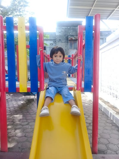 The image presents a young child positioned on a yellow slide within an outdoor playground in Pantegajah, Indonesia. The child, who appears to be between three and five years old, has short, straight, dark hair and dark eyes, and is looking directly at the camera with a neutral expression. The child is dressed in a light blue long-sleeved fleece top with a small embroidered emblem on the left chest, light blue denim shorts, and white closed-toe sandals with straps.

Both of the child's hands are gripping red vertical support bars on either side of the yellow slide, which is part of a larger playground structure. This structure features prominent elements of red, blue, and yellow painted wood or metal posts and railings. The ground surface beneath and around the playground consists of reddish-brown and grey hexagonal paving stones, showing some patches of dirt and wear.

In the background, an older, grey concrete building with a dark roof and visible barred windows is present on the left side. To the right, a bright white wall with patterned circular cutouts extends along the edge of the play area. Above the playground structure, parts of green tree foliage are visible against a bright, overexposed white sky, indicating strong overhead sunlight. A light-colored corrugated roof structure is also visible high on the right side of the background.