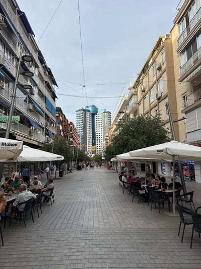 This media file depicts an outdoor seating area of a restaurant or cafe on a paved street in Benidorm, Spain. The scene is set on a bright day, with a clear, light-colored sky suggesting it is daytime. Buildings line the street, with some featuring balconies and awnings. A prominent, tall modern building with a glass facade can be seen in the background.

Several people are seated at tables, engaged in conversations and enjoying their time outdoors. Some are dining, while others appear to be socializing. The overall atmosphere seems relaxed and pleasant. The seating area is furnished with numerous black chairs and white umbrellas providing shade.

Visible text includes "Mahou" printed on the umbrellas, a Spanish beer brand, and "DESDE 1890" also on the umbrellas. Another sign reads "LOUNGE & BAR." There's also text on a green sign that is partially visible. The cobblestone pavement and the architectural style of the buildings are characteristic of a European urban setting. The image appears to be taken from a slightly elevated perspective, looking down the street.