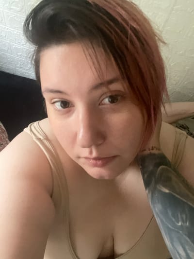 The image shows a close-up selfie of a person lying down. The person has short, dark hair with pinkish undertones, light skin, and brown eyes. They are wearing a light-colored tank top. A tattoo is visible on their arm. The background appears to be a wall with a textured pattern, possibly wallpaper, and a dark surface which could be furniture. The lighting is soft and diffused. The image does not contain information that would allow for the specific location in Korobitsino, Russia to be determined.