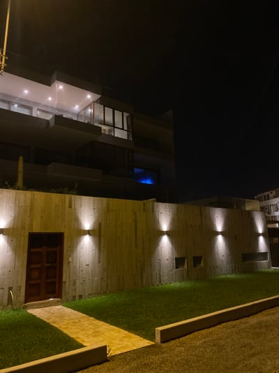 The image depicts a modern building with a concrete exterior wall in the Punta Hermosa district of Lima, Peru. The coordinates provided (latitude: -12.335, longitude: -76.829) correspond to the city of Punta Hermosa. The wall is illuminated by four spotlights, casting a warm glow on the textured surface. A wooden door, slightly ajar, is set into the wall. A small grassy area extends in front of the wall, bordered by a low concrete retaining wall. A paved walkway leads from the door toward the viewer. The top of the building is visible above the wall, with several windows illuminated from within. The building appears to be part of a larger complex, with the rooflines of other structures visible in the distance. The image was taken at night, and the sky is dark, except for a faint glow from streetlights and the building's interior lights.
