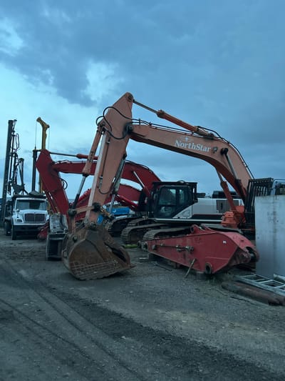 The image shows a lot of heavy construction equipment parked on a gravel lot. There are several excavators, a large truck with a drill rig mounted on it, and various other parts of construction machinery. The excavators are of different colors, including orange, red, and some with black cabs. The sky above is overcast and appears to be dusk or a cloudy day. One of the excavators has "NorthStar" written on its boom. The scene appears to be an equipment yard or staging area. The location is identified as the city of Aurora, United States of America.