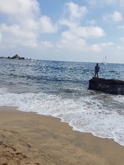 A vertical shot depicts a sandy beach meeting a turquoise ocean. Waves crash onto the shore, creating a white foam border. Dark rocks jut out into the water, and a figure stands atop one of them. In the distance, a sailboat is visible on the horizon. A blue sky dotted with fluffy white clouds completes the serene coastal scene.