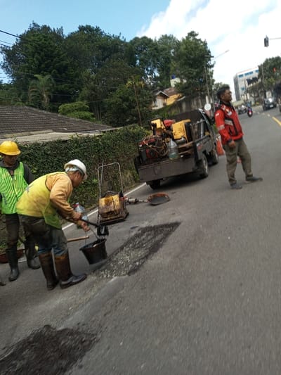 The scene depicts roadside maintenance operations on an asphalt road in the city of Boommlog, Bhutan. Three adult males are present. In the foreground, a male worker wearing a yellow hard hat, light green reflective vest, dark shirt, pants, and dark boots is actively pouring a dark, viscous substance, likely tar or asphalt, from a black bucket onto a patched area of the road using a long-handled tool. To his left, another male worker in a yellow hard hat, bright green reflective vest, dark shirt, pants, and dark boots stands observing. Behind the pouring worker, a yellow and black vibrating plate compactor is positioned on the road, with a dark shovel lying nearby.
A dark-colored pickup truck is parked on the right shoulder of the road, facing away from the camera. Its bed is loaded with machinery, including yellow and red power equipment and a large clear plastic water bottle. To the right of the truck, a third male in a red jacket with reflective strips, dark pants, and dark shoes stands upright, facing towards the right side of the frame, appearing to monitor the surroundings or traffic.
The road surface exhibits dark patches consistent with ongoing repair work. To the left of the road, a low wall covered in dense green foliage separates the road from a building with a dark tiled roof, set against a backdrop of numerous tall green trees. In the distance to the right, other vehicles, including cars and a motorcycle, are visible on the road, along with a traffic light fixture and a multi-story building featuring blue elements. The sky is partly cloudy blue, and daylight illuminates the scene, casting discernible shadows on the road.