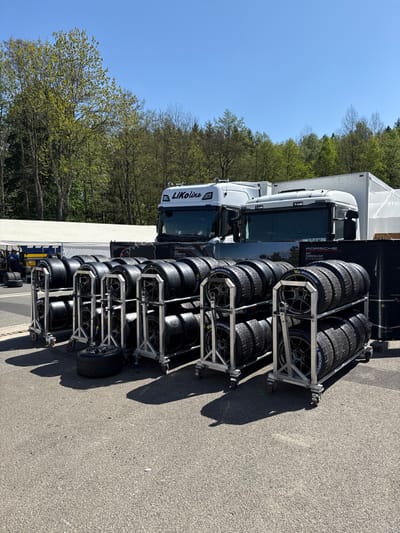 The image depicts a row of tire racks with black tires stacked on them in front of two white semi-trailer trucks. The trucks are parked near a white tent and a black sign with white text on it. The trucks have "LiKoline" written on the side of the cabs. The scene is located in Stavelot, Belgium, at latitude 50.441 and longitude 5.969, and is likely a pit lane or a paddock area at a race track. The tire racks are made of metal and are on wheels. There are green trees in the background. The ground is asphalt and the sky is blue with some clouds. The sun is shining brightly and there are shadows cast by the trucks and the tire racks. There are no people in the image.