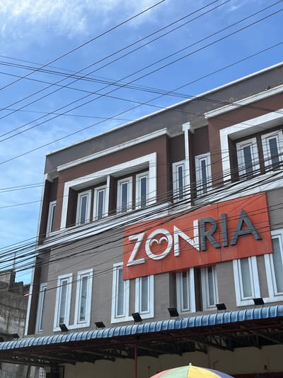 This media file shows the exterior of a multi-story building against a bright blue sky with scattered white clouds. The building has a brown facade with white trim around rectangular windows. A prominent orange sign with large gray letters spells out "ZONRIA," with a stylized white heart shape at the end of the word. Numerous black electrical wires are strung across the frame, obscuring parts of the building and the sky. To the right, there's a section of the building with a blue and white striped awning and some metal framing, and a colorful circular object, possibly an umbrella, is partially visible on the far right. The image appears to be taken during the daytime, under fair weather conditions. The setting suggests an urban or semi-urban environment, given the building architecture and the abundance of overhead wires. The angle of the shot, looking upwards at the building, creates a sense of scale and emphasizes the height. There are no people or specific activities depicted. The visible text is "ZONRIA". The location context provided is Kota Kuala Simpang, Indonesia.