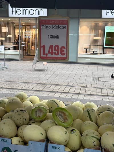 The image shows a street scene in Witten, Germany, with a market stall selling melons in the foreground. A sign above the melons advertises "TOP ANGEBOT Dino Melone 1. Stück 1,49€ WITTEN MARKT". Behind the market stall, a Fielmann optician store is visible with a large illuminated sign. The street is paved with cobblestones, and tram tracks are present. In the background, other shop windows and storefronts can be seen. A pigeon is visible on the right side of the image. The melons are pale yellow with some green speckles and stripes, and some are cut open, revealing their green flesh and seeds. The overall scene depicts a vibrant urban market environment in Witten.